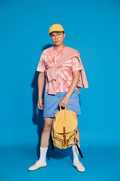 A trendy young man in a pink shirt and blue shorts poses with a yellow backpack against a vibrant blue backdrop. — Stock Photo