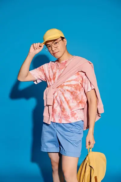 Um jovem elegante posa em um pano de fundo azul, vestindo uma camisa rosa e shorts azuis enquanto segura um saco amarelo. — Fotografia de Stock