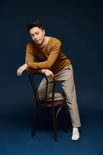 Un joven de moda con un atuendo elegante se sienta con confianza en la parte superior de una silla de madera, golpeando una pose dinámica. - foto de stock