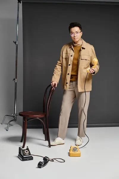 Un joven de moda está de pie con confianza junto a una silla, sosteniendo el teléfono - foto de stock