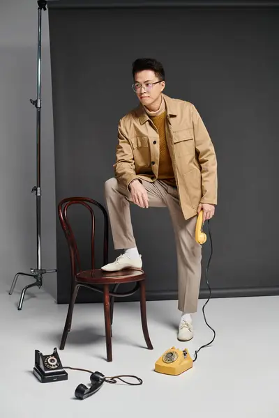 Un joven de moda con un atuendo elegante sentado en la parte superior de una silla al lado del teléfono, exudando confianza y carisma. - foto de stock