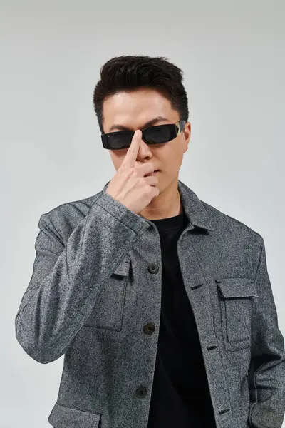 Um jovem elegante faz uma pose confiante em uma jaqueta cinza elegante e óculos de sol na moda. — Fotografia de Stock