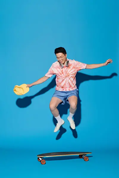 A stylish young man jumps gracefully in the air while holding a skateboard, exuding energy and style on a blue backdrop. — Stock Photo