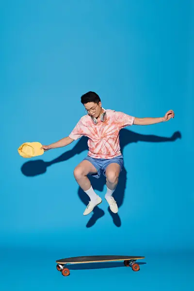 Un joven elegante y guapo con un atuendo moderno salta en el aire con un monopatín sobre un fondo azul. - foto de stock