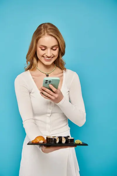 Une femme blonde tient gracieusement un plateau de sushi dans une main et un téléphone portable dans l'autre, incarnant l'élégance multitâche. — Photo de stock