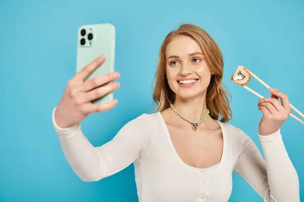 Стильная женщина с светлыми волосами, держащая суши и палочки в руке и сотовый телефон в другой. — стоковое фото
