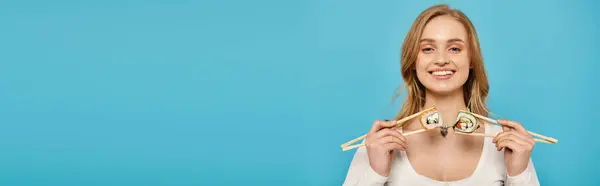 A glamorous woman with blonde hair holding two chopsticks with sushi perched on them in front of her face. — Stock Photo