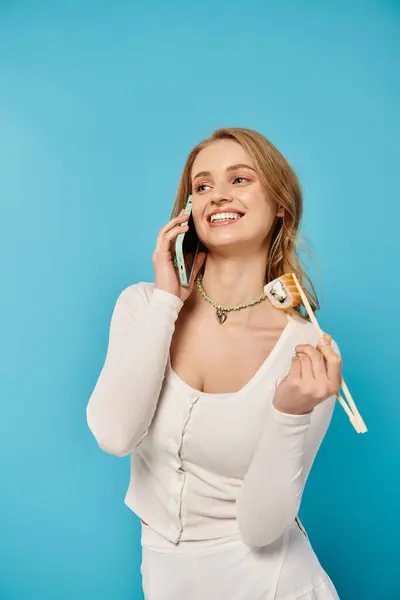 Une femme gracieuse en robe blanche engage une conversation téléphonique tenant des baguettes avec des sushis. — Photo de stock