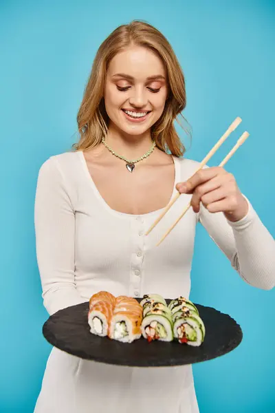 Femme blonde tient gracieusement assiette de sushi et baguettes, prêt à se livrer à un délicieux repas asiatique. — Photo de stock