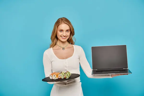 Una hermosa mujer rubia posa elegantemente con un plato de delicioso sushi en la mano y un portátil en otro. - foto de stock