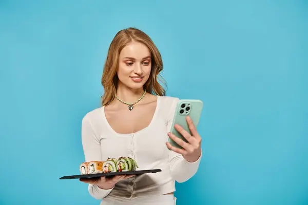 Una mujer elegante con el pelo rubio sosteniendo un plato de sushi y un teléfono celular, golpeando una pose. - foto de stock