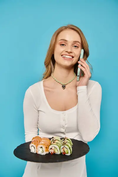 Una hermosa mujer rubia posando mientras sostiene un plato lleno de sushi en una mano y un teléfono celular en la otra. - foto de stock