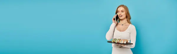 Una mujer rubia impresionante sostiene una bandeja de sushi mientras charla en su teléfono celular. - foto de stock