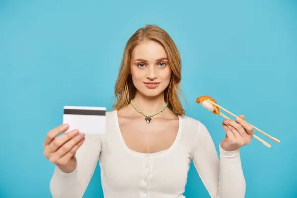Una mujer rubia sostiene con confianza una tarjeta de crédito mientras muestra un delicioso sushi asiático. - foto de stock