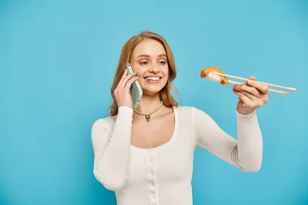 Une femme blonde tient délicatement des baguettes tout en bavardant au téléphone, appréciant la cuisine asiatique. — Photo de stock
