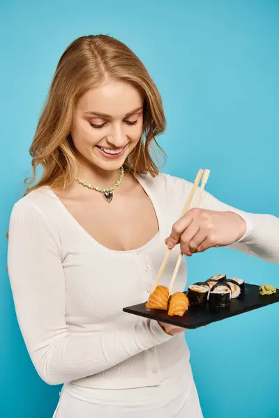 Una hermosa mujer con el pelo rubio delicadamente sostiene un plato de sushi y palillos. - foto de stock