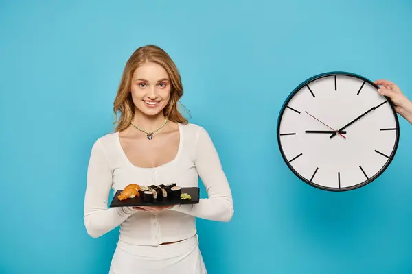 Eine blonde Frau hält einen Teller mit asiatischem Essen vor einer Uhr. — Stockfoto
