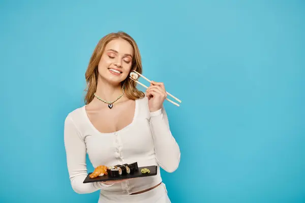 Шикарная блондинка держит поднос суши и палочек для еды, готовая насладиться восхитительным азиатским кулинарным опытом. — стоковое фото