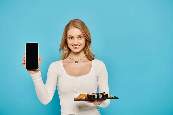 Una mujer rubia elegante elegantemente multitareas, sosteniendo un plato de cocina asiática en una mano y un teléfono celular en la otra. - foto de stock