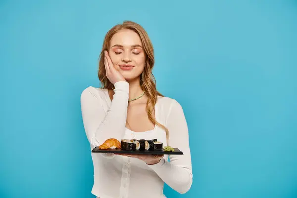 A blonde woman gracefully holds a tray of sushi against a serene blue background. — Stock Photo