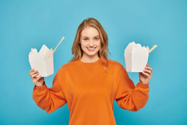Une femme aux cheveux blonds tenant deux boîtes de nourriture asiatique dans ses mains, à la recherche coûteuse et contemplative. — Photo de stock