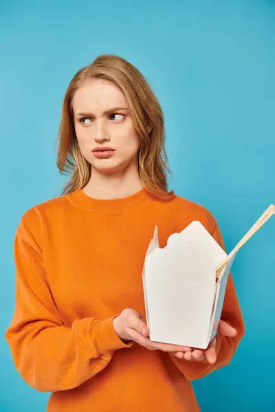 Una mujer elegante en un suéter naranja sostiene con gracia una deliciosa caja de comida, mostrando una mezcla de moda y gastronomía. - foto de stock