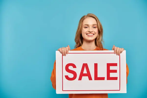A blonde woman confidently holds a sign that reads Sale against a soft background. — Stock Photo