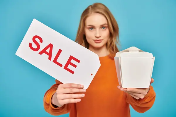 A beautiful blonde woman energetically holds a sale sign and a box of delicious Asian food while showcasing her products. — Stock Photo