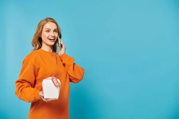 Una mujer con estilo en un suéter naranja se involucra en una animada conversación en un teléfono celular con caja de comida en la mano. - foto de stock