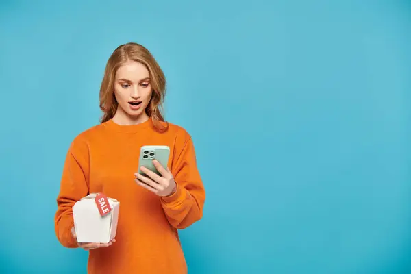 Una mujer con estilo en un suéter naranja sostiene un teléfono celular con confianza y comida boz woth etiqueta de ventas en él. - foto de stock