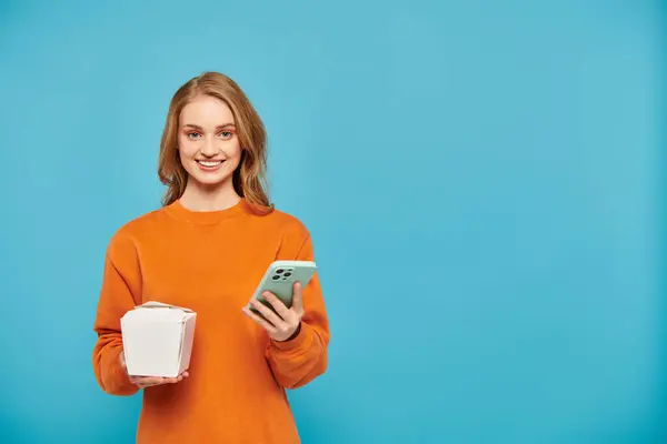 Una mujer con cabello rubio sosteniendo la caja de comida y un teléfono celular mientras está en el telón de fondo azul - foto de stock