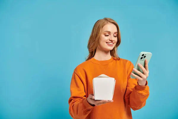 Une femme blonde équilibre sans effort une boîte de nourriture asiatique dans une main tout en faisant défiler son téléphone portable avec l'autre. — Photo de stock
