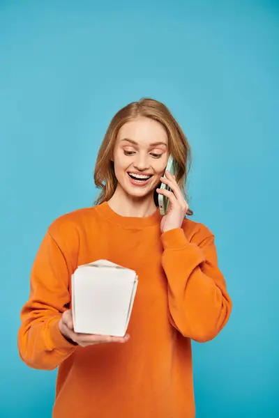 Une femme élégante aux cheveux blonds tient une boîte de nourriture asiatique tout en bavardant sur son téléphone portable. — Photo de stock