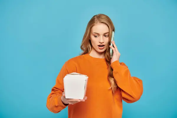 Una mujer con estilo en un suéter naranja está hablando animadamente en un teléfono celular y mirando la caja de comida. - foto de stock