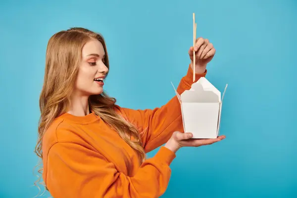 Eine stilvolle Frau in orangefarbenem Hemd hält selbstbewusst eine Schachtel mit asiatischem Essen und Essstäbchen in der Hand.. — Stockfoto
