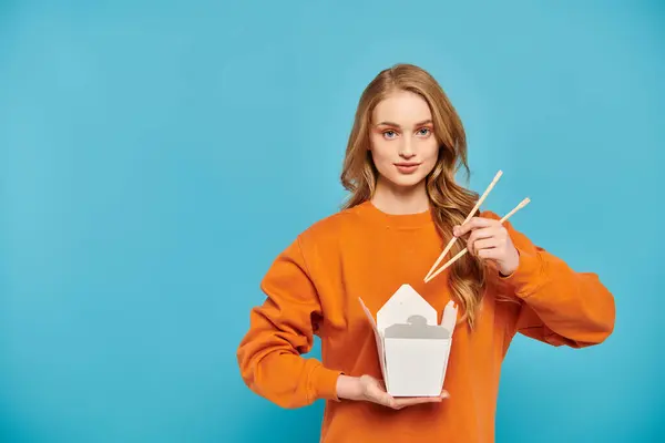 A beautiful blonde woman delicately holds chopsticks and a box of delicious Asian food. — Stock Photo