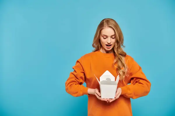 A beautiful blonde woman in an orange sweater joyfully holding a box of delicious Asian food. — Stock Photo