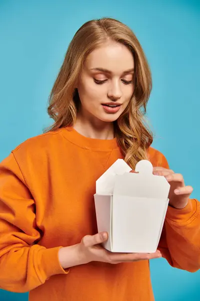 Una bella donna in un maglione arancione tiene una scatola di cibo bianco, la sua espressione curiosa e deliziata. — Foto stock