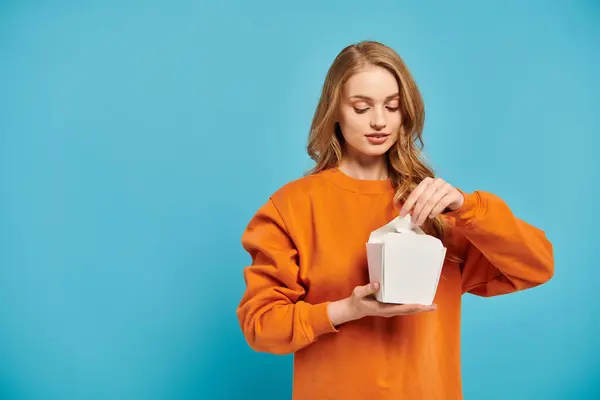 Eine schöne blonde Frau im orangefarbenen Pullover hält eine weiße Lebensmittelbox in der Hand. — Stockfoto