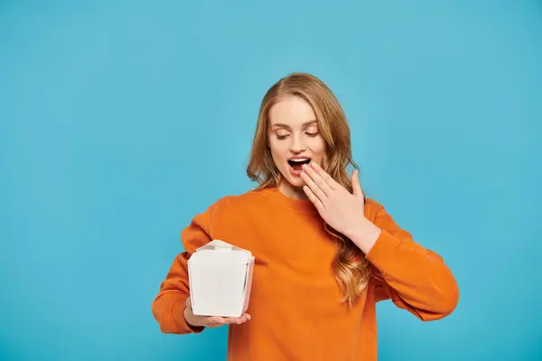 A woman with blonde hair holding a box of Asian food in front of her face. — Stock Photo