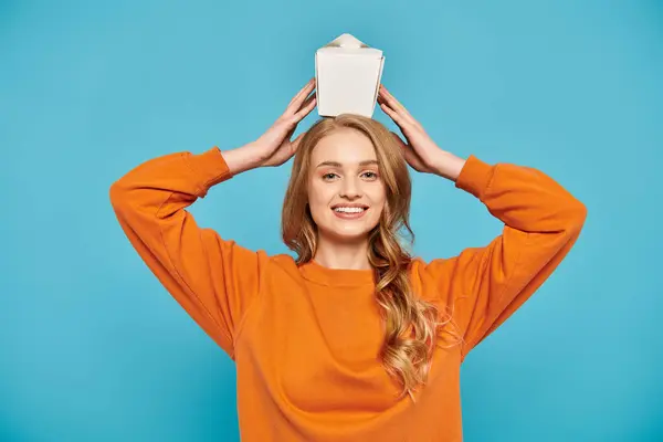 A beautiful woman in an orange sweater joyfully holds box of Asian food over her head. — Stock Photo