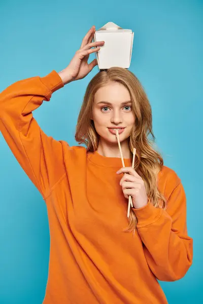 A stylish woman in an orange sweater confidently holds box of Asian food and chopsticks. — Stock Photo