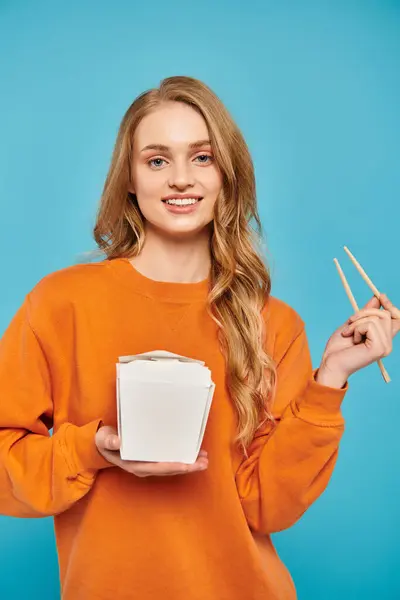 Une belle femme aux cheveux blonds tient délicatement boîte à nourriture et baguettes, savourant la cuisine asiatique. — Photo de stock