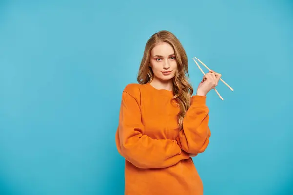 A beautiful woman in an orange sweater delicately holds a pair of chopsticks, ready to enjoy a delicious Asian meal. — Stock Photo