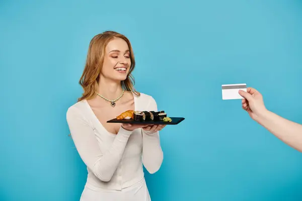 Une superbe femme blonde dans une robe blanche immaculée tient une assiette de nourriture asiatique appétissante, main avec carte de crédit. — Photo de stock