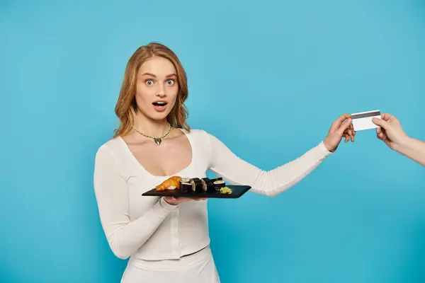 Eine schöne blonde Frau in einem weißen Kleid hält elegant ein Tablett mit köstlicher asiatischer Küche in der Hand, Hand mit Kreditkarte. — Stockfoto