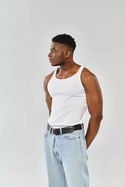 A young, sexy African American man showcases his toned physique in a white tank top and jeans against a grey studio backdrop. — Stock Photo
