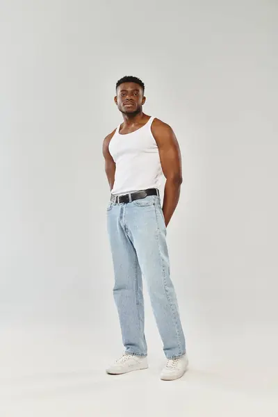 A young and sexy African American man confidently poses in a white tank top and jeans against a grey studio backdrop. — Stock Photo