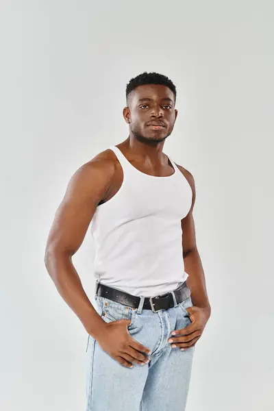 A young, sexy African American man in a white shirt and jeans strikes a pose in a studio against a grey background. — Stock Photo