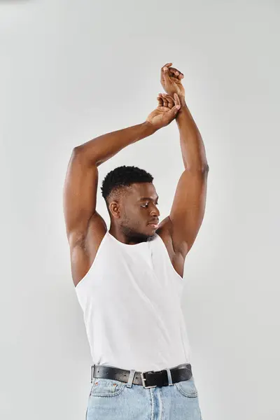 Um jovem afro-americano em um top branco, levantando as mãos no ar em um estúdio contra um fundo cinza. — Fotografia de Stock